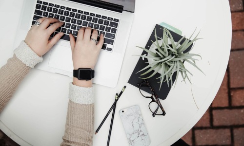 Find your perfect fit at 610 West overhead shot of woman working on a laptop at a round table with pencils, her cell phone, glasses, notebook and a potted plant